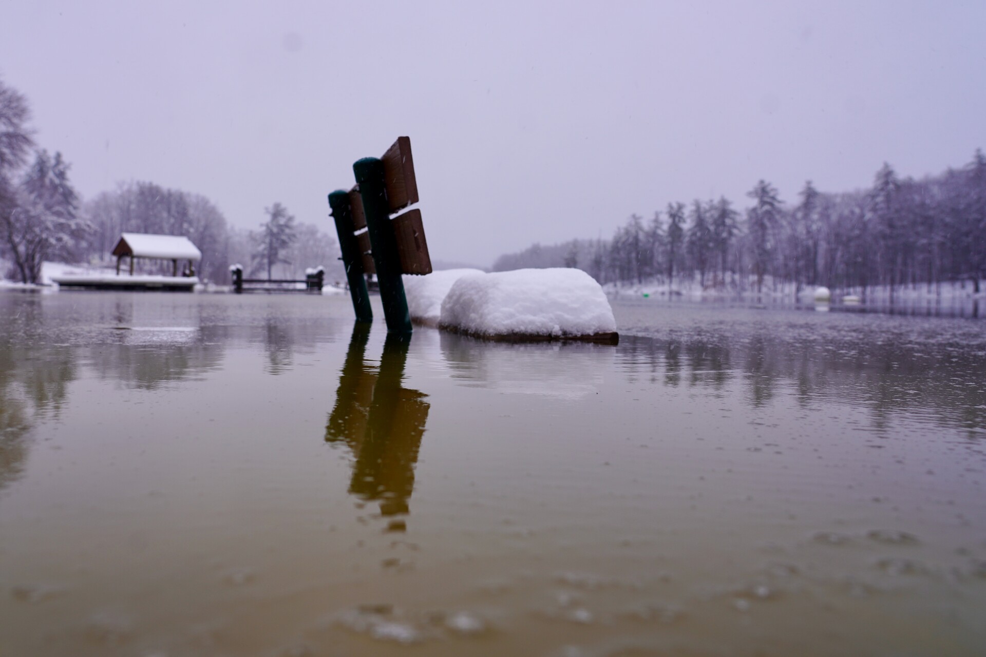 Grey sky. The camera is held at water level, which rises to the level of a seat on the park bench. Snow covers the park bench and trees in the background.