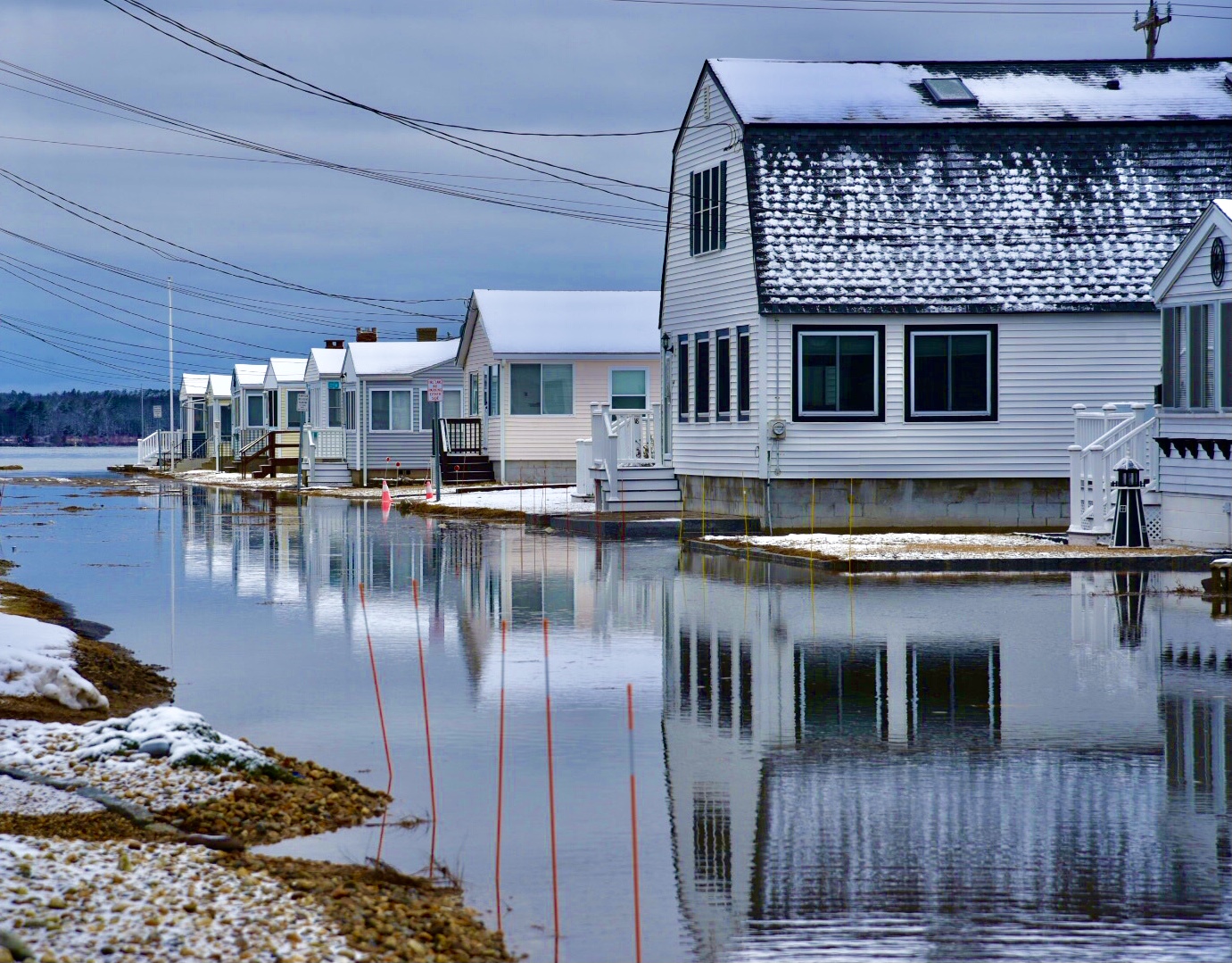 A row of homes reflected into the water on the road in front of them. Snow covers their roofs.