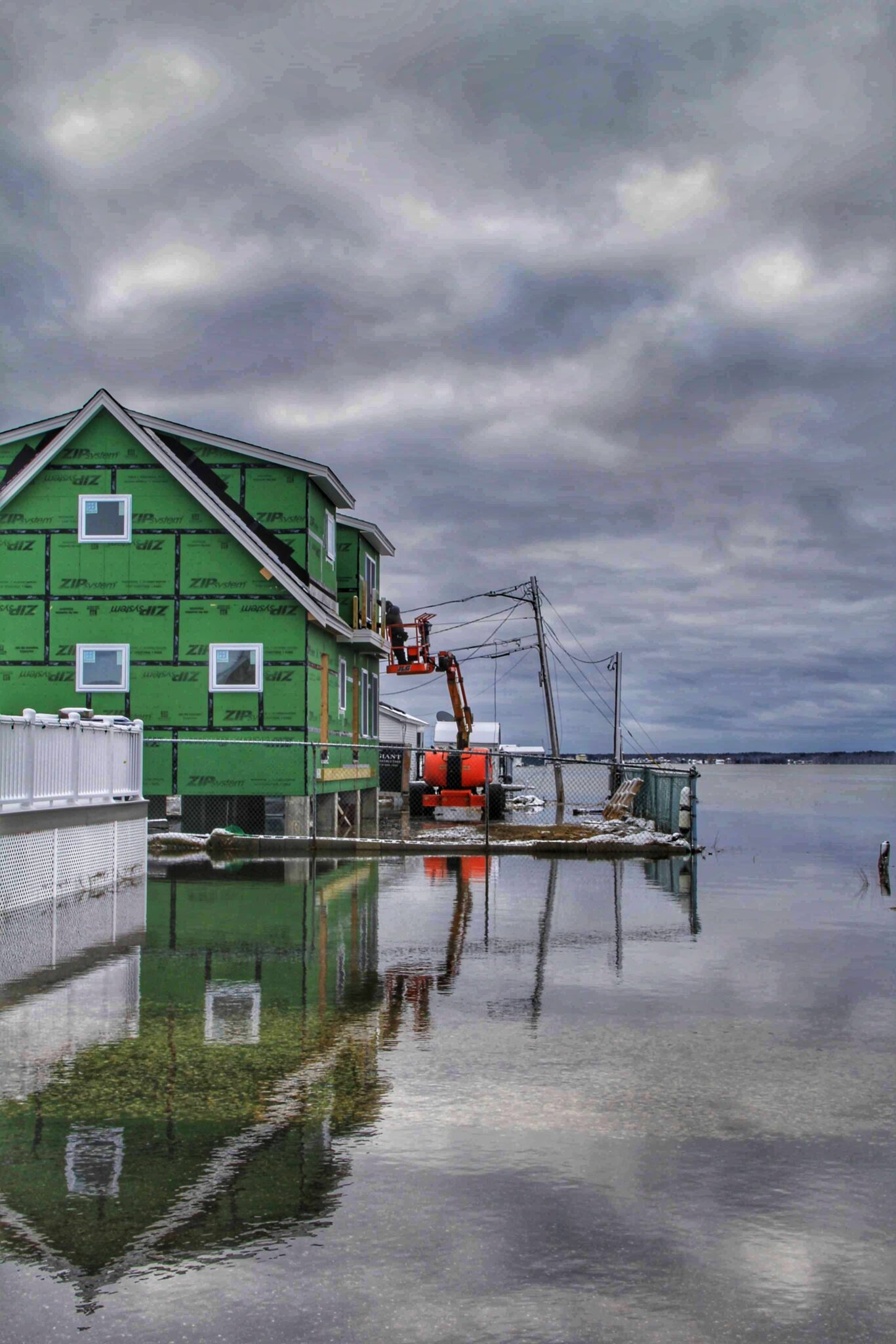 Cloudy sky and flooded street. A new house with a chain link fence around it is being built. The house is elevated on above the flooded water.