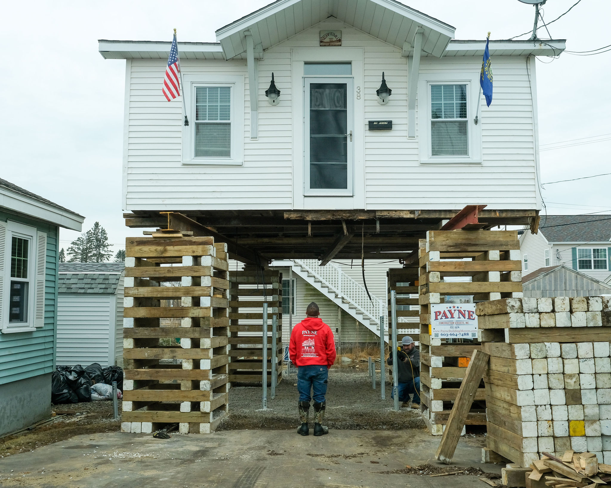 A man in a red sweatshirt is looking up at a white cottage elevated on wood pilings. 