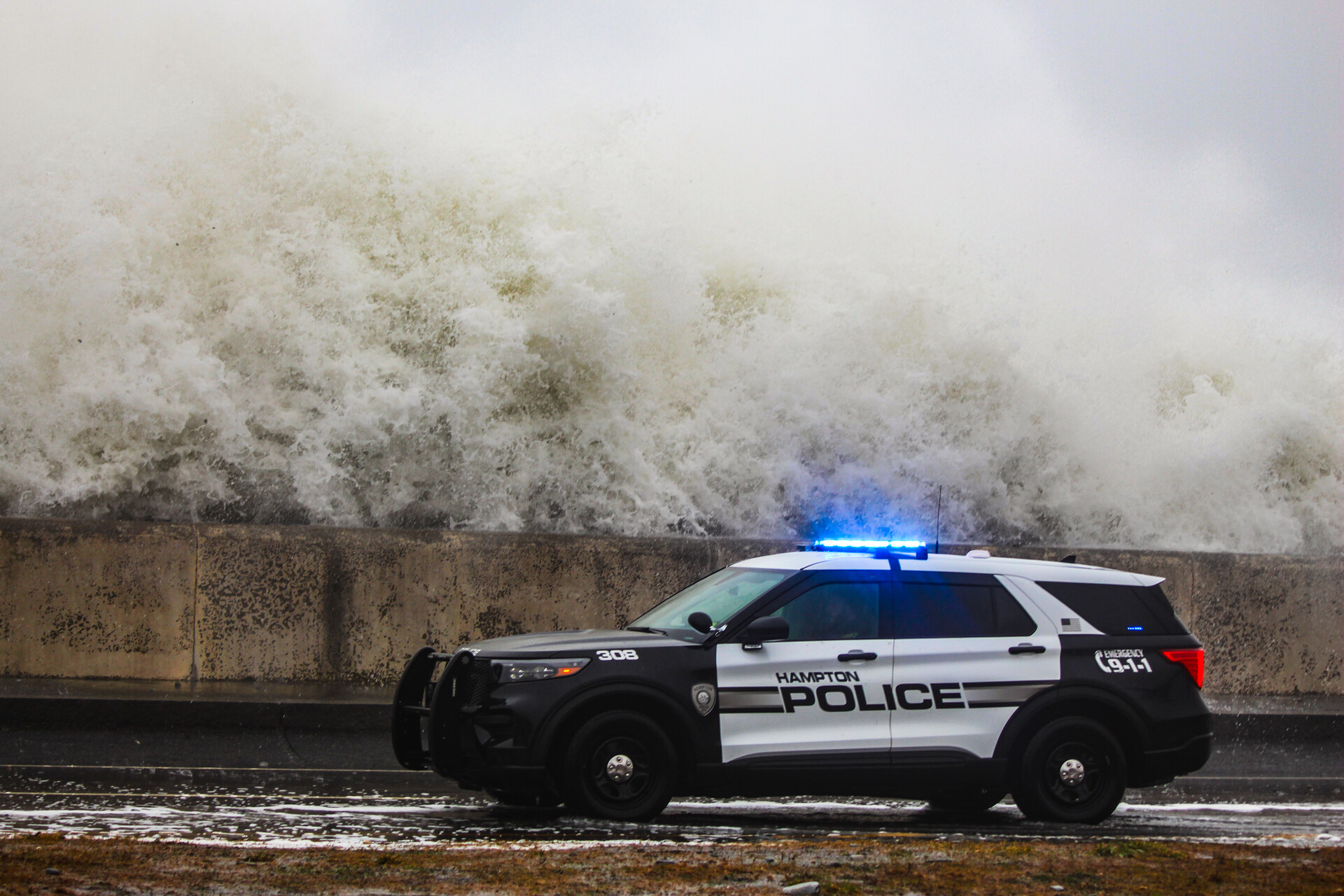 A police car with its lights on drives in front of a sea wall with foaming white waves crashing over the wall.