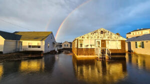 Rainbow in a cloudy blue sky over several cottages that are surrounded by flood water.