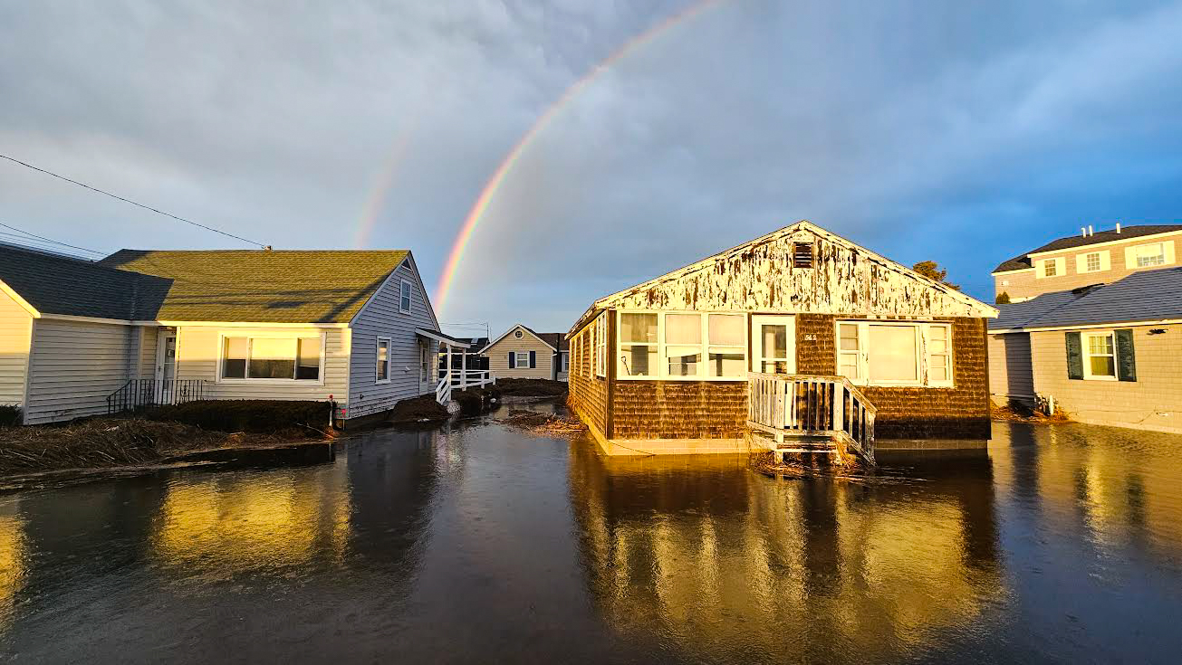 Rainbow in a cloudy blue sky over several cottages that are surrounded by flood water.