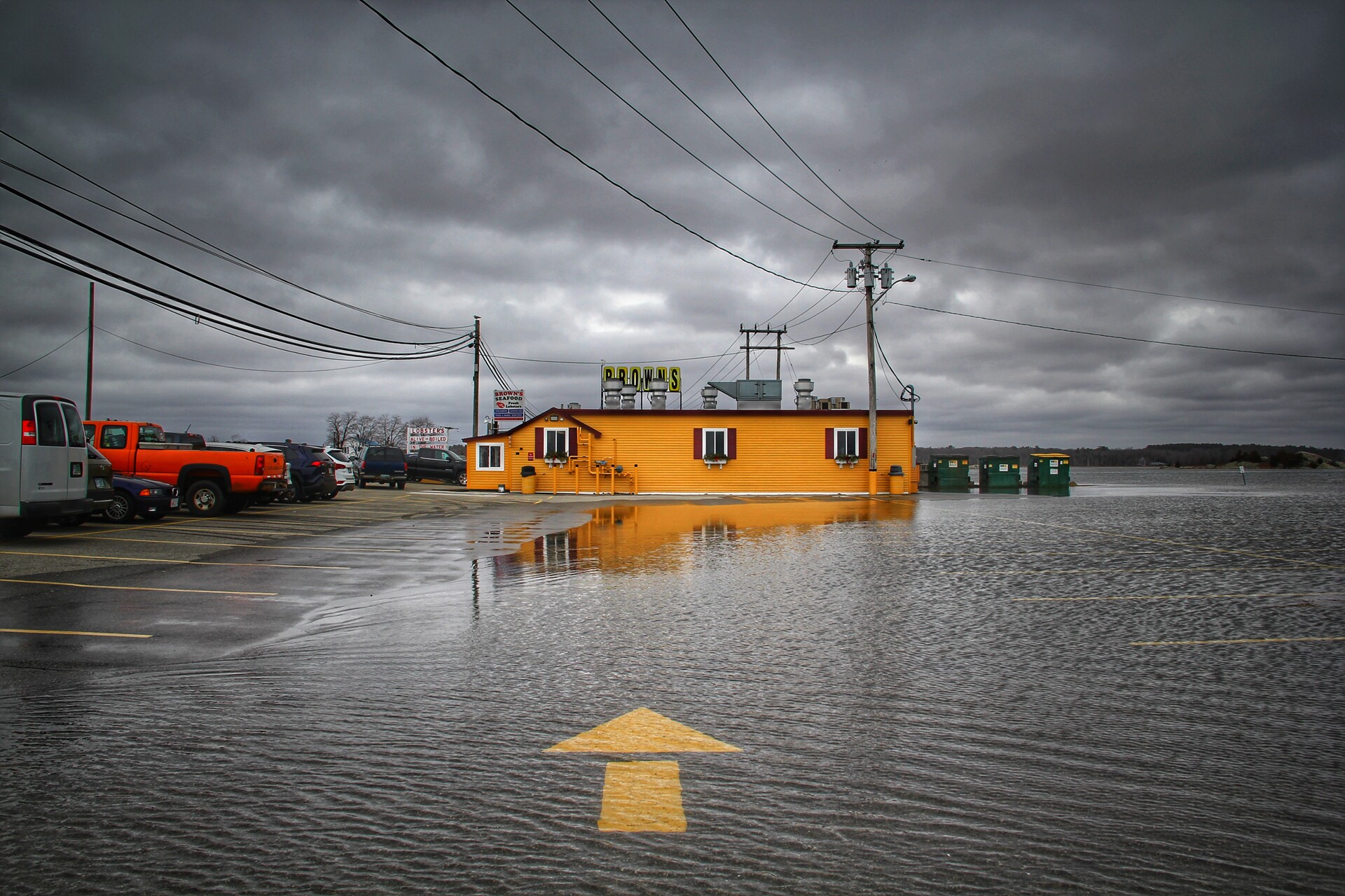 A grey cloudy sky over a bright yellow building surrounded by a flooded parking lot with a big yellow painted arrow.