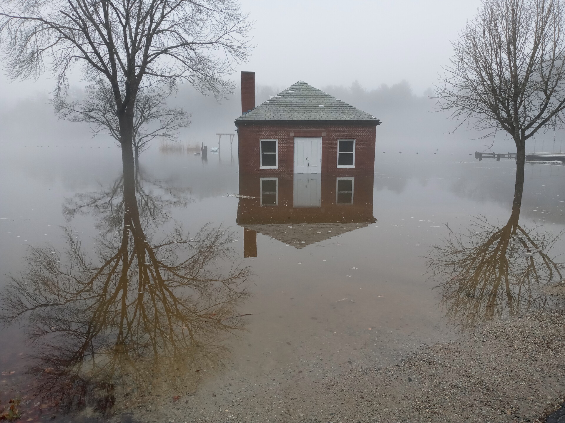 A foggy day. A small brick building is surrounding by flood water. Its reflection and that of a nearby tree are visible in the water.
