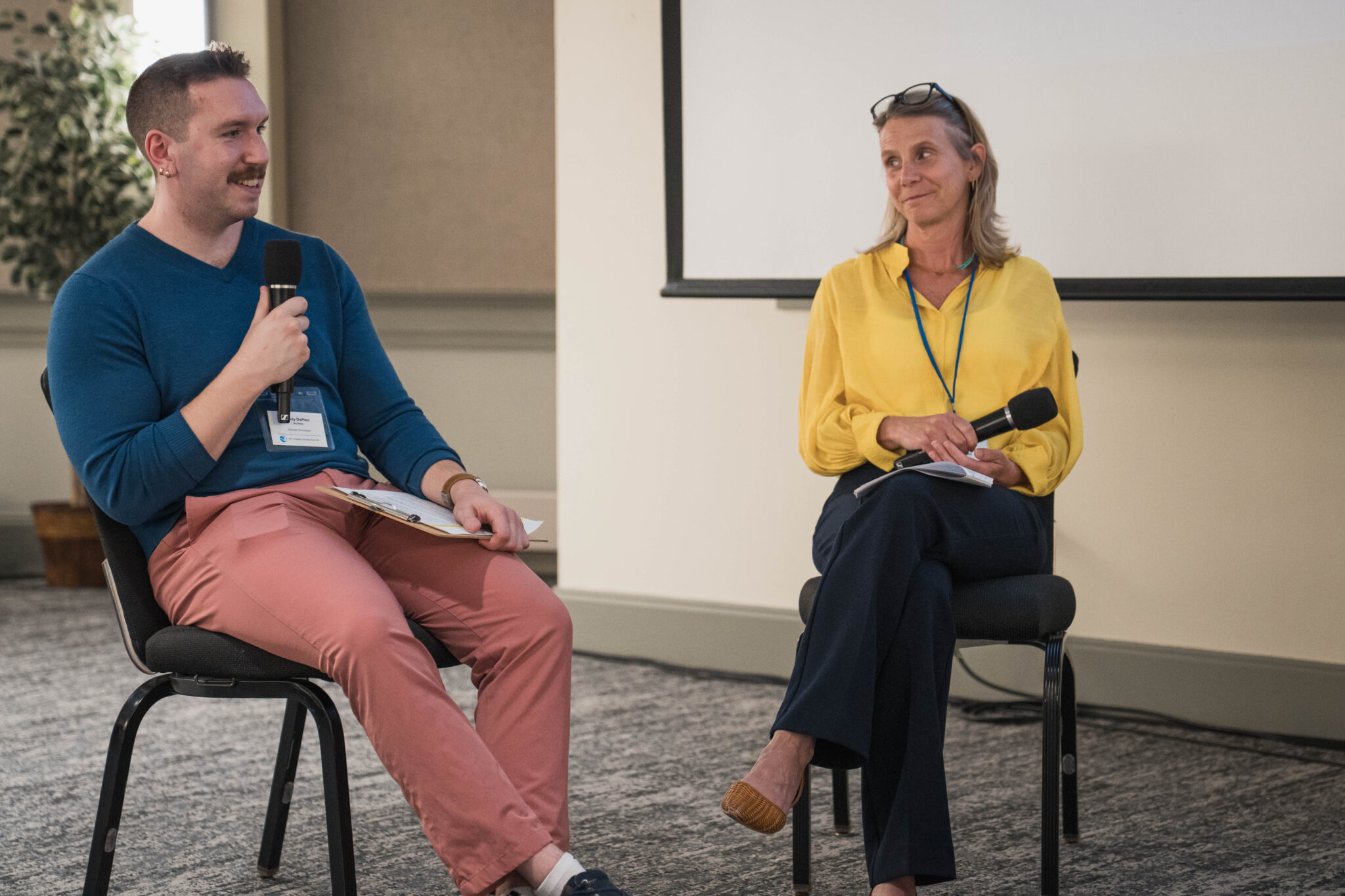 A man and a woman on a conference panel smile at one another.