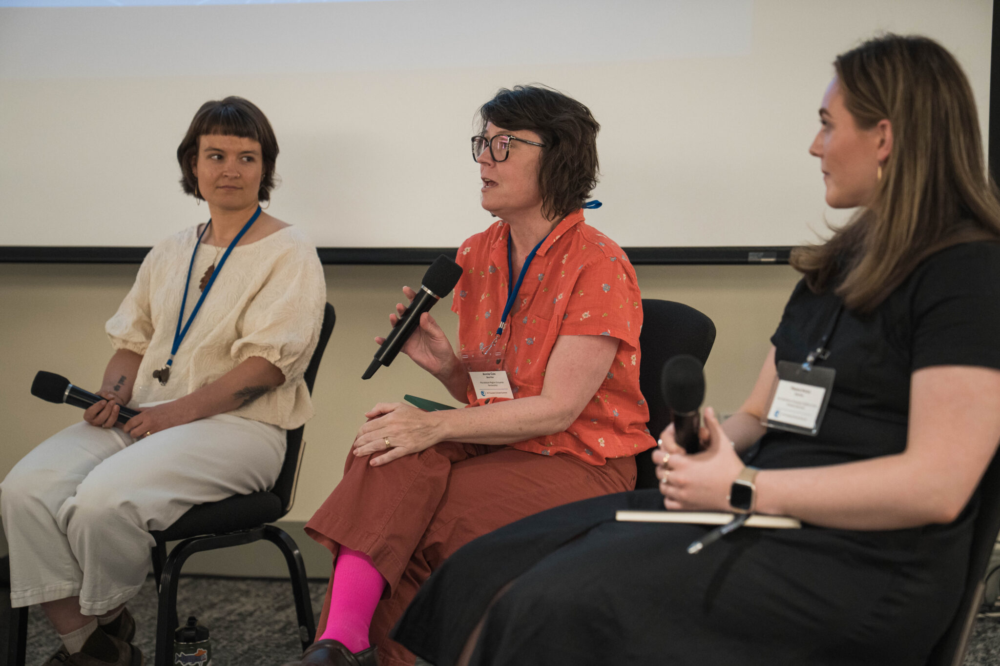 A row of three women on a conference panel, the middle one speaks into a mic